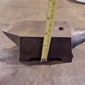 May include: A small, rusty metal anvil with a blue-painted horn. The anvil is being measured with a tape measure that reads 7 inches.