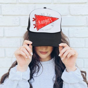 Pode incluir: Bon&eacute; de caminhoneiro preto e branco com uma fl&acirc;mula vermelha que diz "Rangers". O bon&eacute; tem uma parte traseira de malha e uma aba curva. O texto "Trading Company" est&aacute; impresso no bon&eacute;.