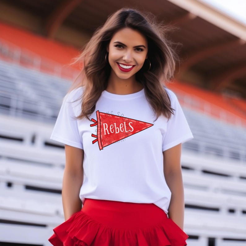 Pode incluir: Camiseta branca com um design de fl&acirc;mula vermelha que diz "Rebels". A fl&acirc;mula tem formato triangular com fundo vermelho e letras brancas. O look &eacute; complementado por uma saia vermelha.