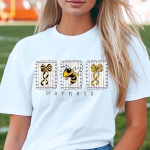 May include: White t-shirt featuring three square designs with a bee and bows. The center square has a yellow and black bee. The other squares have bows with black and yellow stripes. The text "Hornets Trading Company" is below the designs.