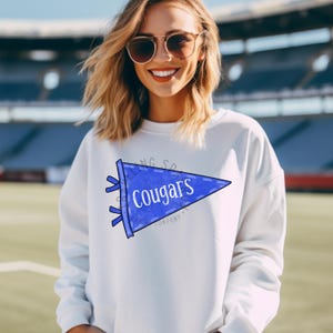 Pode incluir: Camisola branca com um gr&aacute;fico de fl&acirc;mula azul que diz "Cougars" em texto branco. A fl&acirc;mula tem uma borda azul e fitas. A camisola &eacute; de estilo casual.