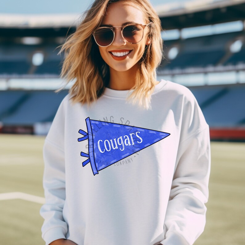 Pode incluir: Camisola branca com um gr&aacute;fico de fl&acirc;mula azul que diz "Cougars" em texto branco. A fl&acirc;mula tem uma borda azul e fitas. A camisola &eacute; de estilo casual.