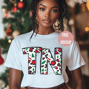 Puede incluir: Camiseta blanca con un contorno negro de las letras "TN" rellenas con un estampado de leopardo con árboles de Navidad rojos, verdes y blancos y estrellas. Maqueta gratuita.