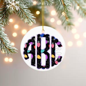 Puede incluir: Adorno navideño redondo de cerámica blanca con una cuerda dorada para colgar. El adorno presenta las letras "ABC" en una fuente negra decorativa, delineadas con púrpura. Las letras están rellenas de luces navideñas de colores.