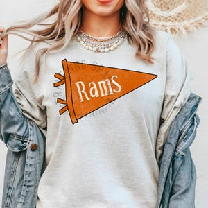 Pode incluir: Camiseta cinza claro com uma fl&acirc;mula laranja com a palavra "Rams" em branco. A fl&acirc;mula tem uma contorno preto e franjas laranja. A camisa tamb&eacute;m tem o texto "TRAV" e "COMPANY".