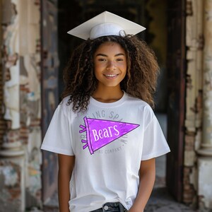 Pode incluir: Uma pessoa vestindo uma camiseta branca com um design de fl&acirc;mula roxa que diz "Bears". A pessoa tamb&eacute;m est&aacute; usando um chap&eacute;u de formatura branco. O fundo &eacute; um ambiente externo desfocado.