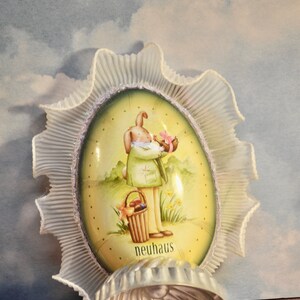 May include: An Easter-themed egg-shaped decoration with a bunny illustration, framed by a ruffled, white trim. A silver-colored, shell-shaped mold sits in front of the decoration. The word "neuhaus" is printed on the egg.