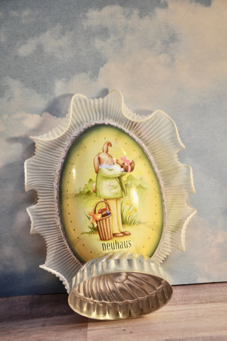 May include: An Easter-themed egg-shaped decoration with a bunny illustration, framed by a ruffled, white trim. A silver-colored, shell-shaped mold sits in front of the decoration. The word "neuhaus" is printed on the egg.