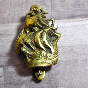 May include: A brass door knocker shaped like a sailing ship with intricate details. The knocker is mounted on a wooden surface.