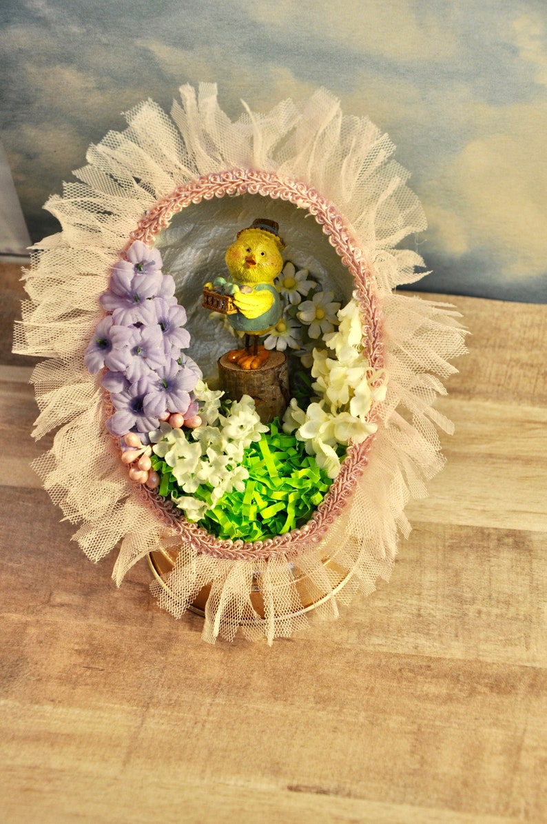 May include: A decorated Easter egg with a pink and white lace trim. Inside the egg is a small yellow chick figurine holding a basket of eggs. The egg is surrounded by white and purple flowers and green grass.