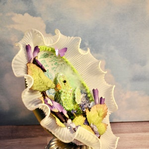 May include: A decorative, whimsical art piece featuring a small, green creature nestled within a ruffled, white fabric structure. The piece is adorned with purple and green floral accents, a small butterfly, and rests on a silver, fluted base.