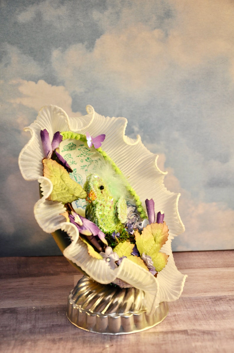 May include: A decorative, whimsical art piece featuring a small, green creature nestled within a ruffled, white fabric structure. The piece is adorned with purple and green floral accents, a small butterfly, and rests on a silver, fluted base.