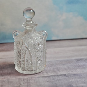 May include: A clear glass bottle with a textured surface and a round stopper. The bottle has two handles on the sides.