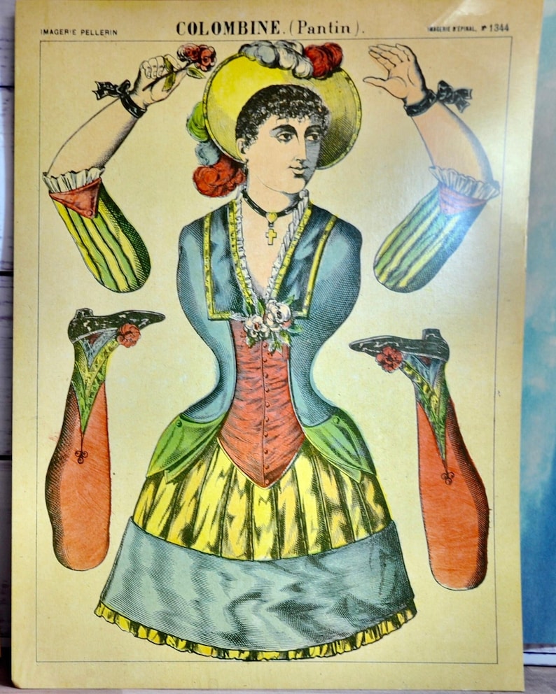 May include: A vintage paper doll sheet titled COLOMBINE (Pantin) featuring a central figure in a blue, red, and yellow dress with a hat, surrounded by detached arms and legs. The illustration includes a cross necklace and floral accents.