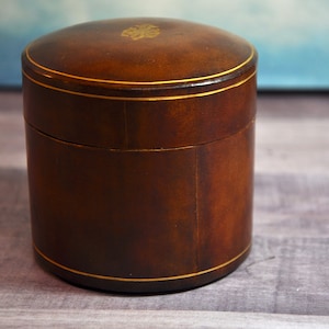 May include: A cylindrical brown leather box with a removable lid. The exterior features thin gold-colored circular lines around the lid and base, and a small gold-colored emblem is centered on the top surface of the lid.