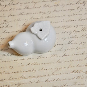 May include: A white ceramic bottle stopper shaped like a gourd with a small leaf design on top. The stopper has a hole in the top for inserting a bottle.