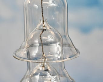 May include: Clear glass bell-shaped decorative hanging ornaments. Each bell has a flared base and a narrower top, suspended by a thin, twisted metal wire. The background is a gradient of blue and white.