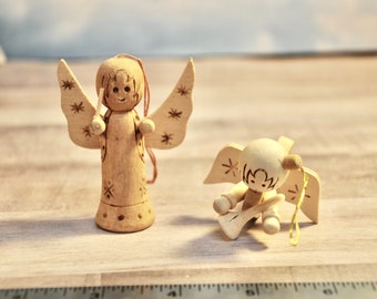 May include: Two wooden angel ornaments. One stands upright with detailed wings and a smiling face. The other is seated, playing a small instrument. Both are carved from light-colored wood, with star and floral accents.