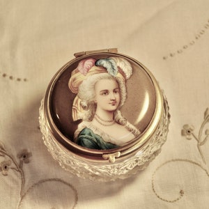 May include: A vintage, round trinket box with a portrait of a woman in a bonnet. The box has a gold-colored rim and a clear, faceted base. The portrait features a woman with white hair, a blue dress, and a pearl necklace.