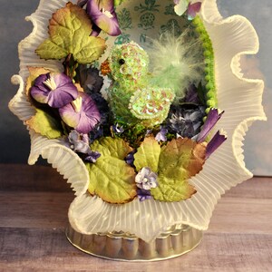 May include: A decorative Easter-themed display featuring a sequined green duckling nestled within a ruffled white lace frame. The arrangement includes purple flowers, green leaves, and a small butterfly, all set on a silver-toned base.