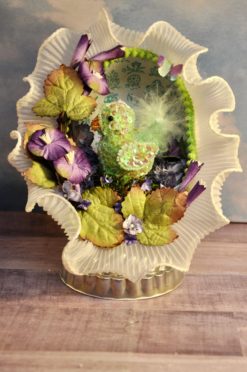May include: A decorative Easter-themed display featuring a sequined green duckling nestled within a ruffled white lace frame. The arrangement includes purple flowers, green leaves, and a small butterfly, all set on a silver-toned base.