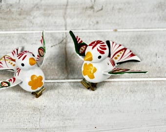 May include: Two white bird ornaments with red, green, and yellow accents. The birds have outstretched wings with red and white patterns. Each bird has a yellow flower shape on its chest and a small gold-colored base.