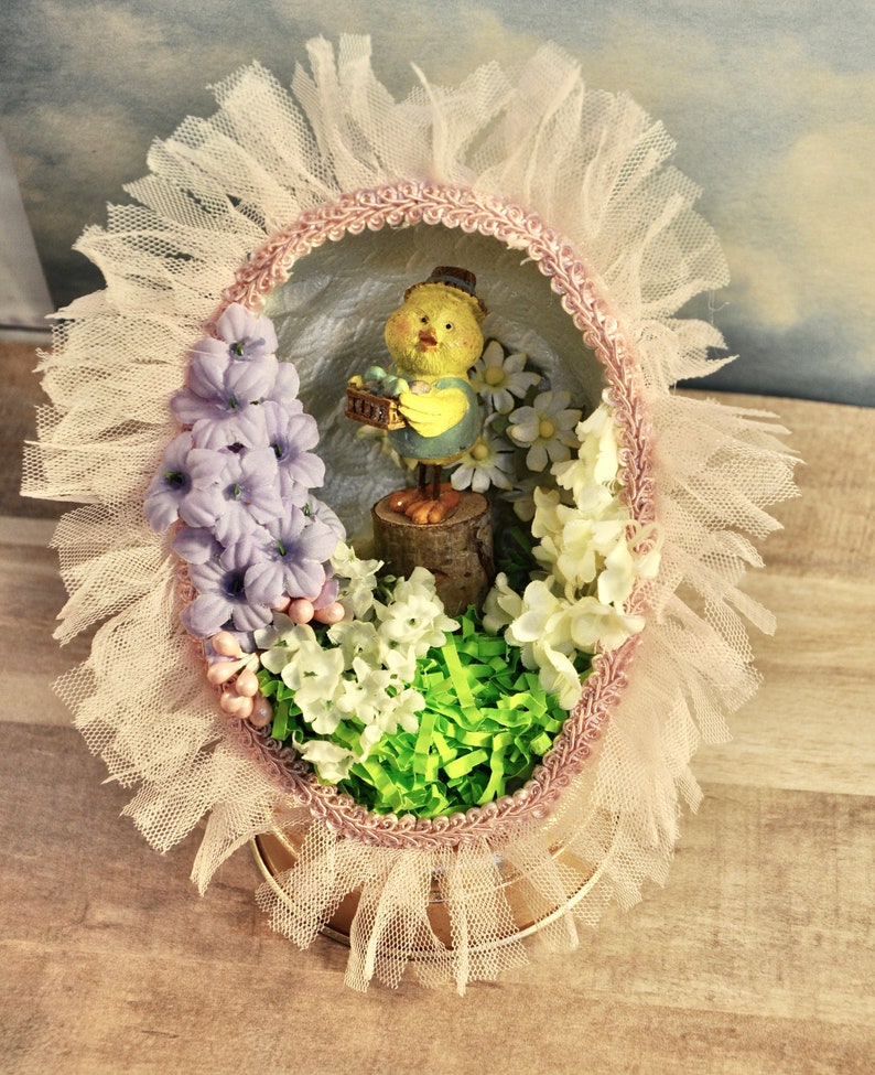 May include: A pink and white Easter egg with a yellow chick figurine holding a basket of eggs. The egg is decorated with lace and flowers.