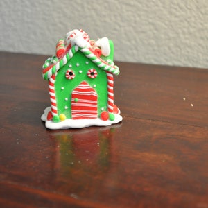 May include: A festive, miniature gingerbread house figurine. The house features a green exterior with red and white striped accents, a red door, and candy cane decorations. The roof is topped with white icing and more candy. A charming holiday decoration.