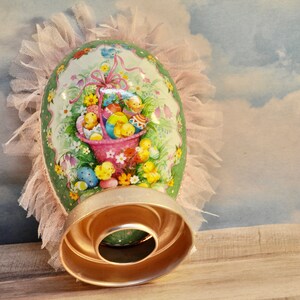 May include: A green and white Easter egg with a pink basket filled with yellow chicks and colorful eggs. The egg is decorated with flowers and is surrounded by a pink tulle ruffle.