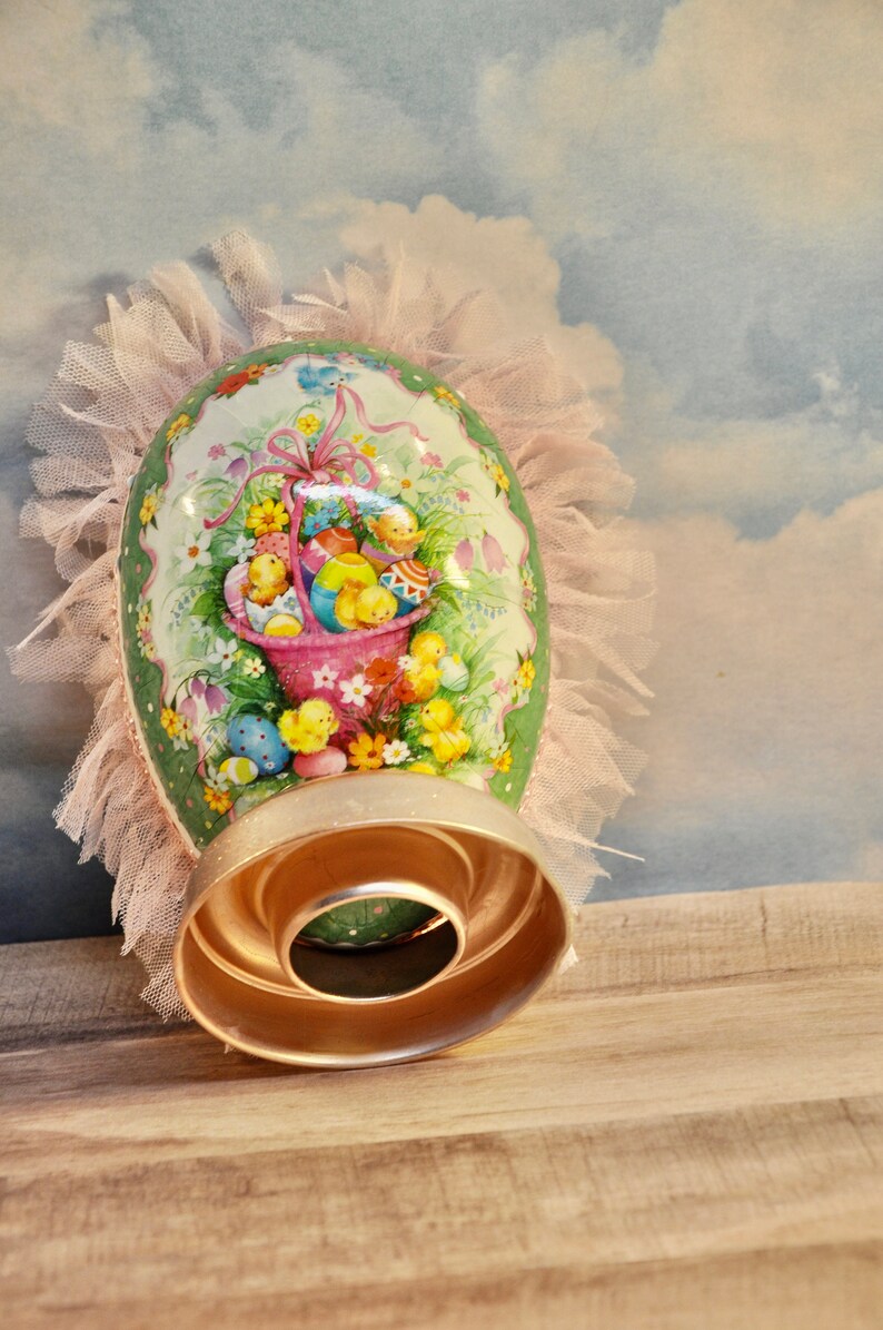 May include: A green and white Easter egg with a pink basket filled with yellow chicks and colorful eggs. The egg is decorated with flowers and is surrounded by a pink tulle ruffle.