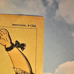 May include: A vintage paper print featuring a detailed illustration of an arm with a dark wristband and a bow, positioned next to the text IMAGERIE D'&Eacute;PINAL, N 1344. The print has a beige background and is set against a blue sky with white clouds.