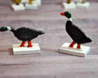 May include: Two black and green painted duck figurines with orange feet stand on white bases. The ducks are facing opposite directions.