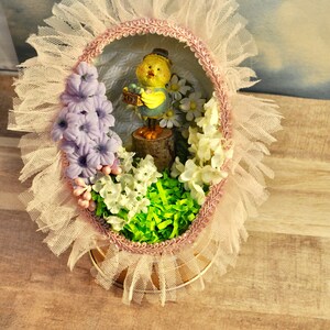 May include: A decorated Easter egg with a pink and white lace trim. Inside the egg is a small figurine of a yellow chick holding a basket of eggs. The egg is surrounded by white and purple flowers and green grass.