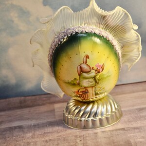 May include: An Easter-themed decorative egg with a painted scene of a bunny holding flowers. The egg is set on a silver-colored base and topped with a ruffled, cream-colored fabric. A decorative item.