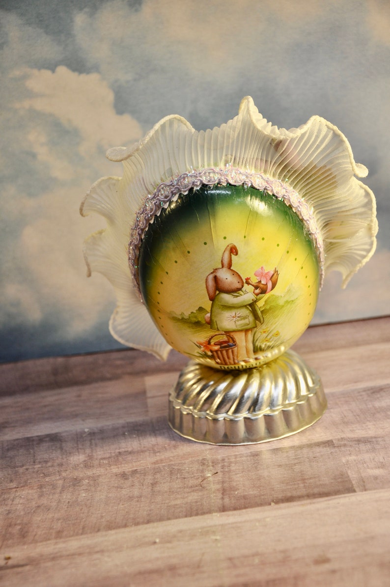 May include: An Easter-themed decorative egg with a painted scene of a bunny holding flowers. The egg is set on a silver-colored base and topped with a ruffled, cream-colored fabric. A decorative item.