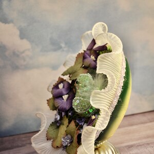 May include: A decorative, egg-shaped ornament with a green and gold base, adorned with purple flowers, green leaves, and a ruffled white trim. The ornament sits on a silver-colored pedestal, creating a unique display piece.