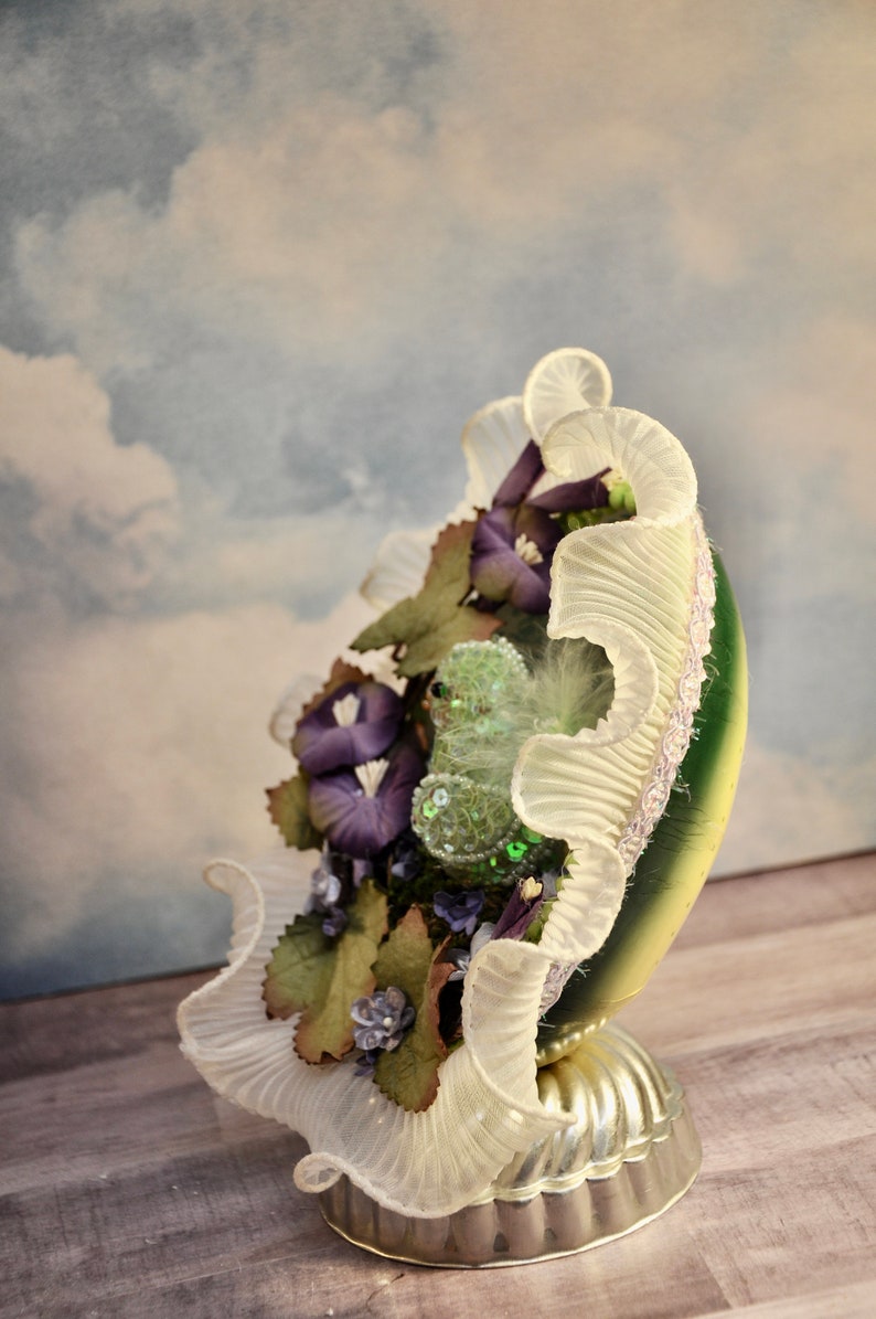 May include: A decorative, egg-shaped ornament with a green and gold base, adorned with purple flowers, green leaves, and a ruffled white trim. The ornament sits on a silver-colored pedestal, creating a unique display piece.