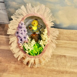 May include: A pastel pink and white Easter egg decoration with a yellow chick figurine holding a basket of eggs. The egg is surrounded by white and purple flowers and green grass.