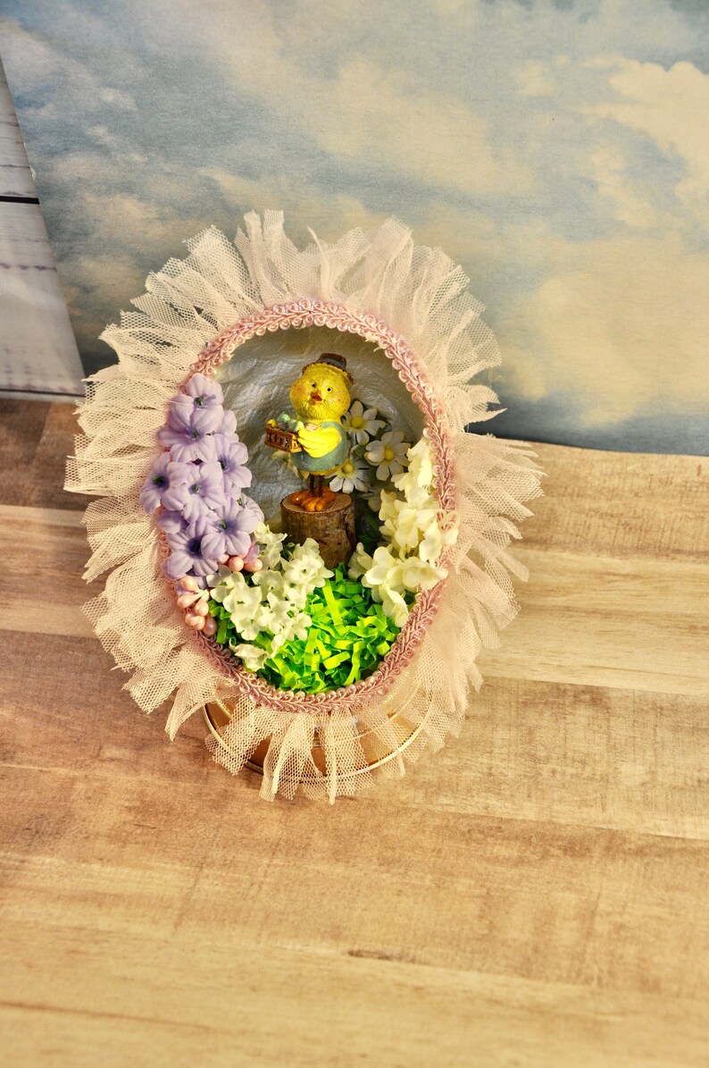 May include: A pastel pink and white Easter egg decoration with a yellow chick figurine holding a basket of eggs. The egg is surrounded by white and purple flowers and green grass.