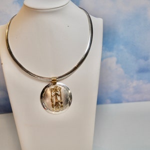 May include: A silver snake chain necklace with a round pendant. The pendant is silver with a gold design in the center.