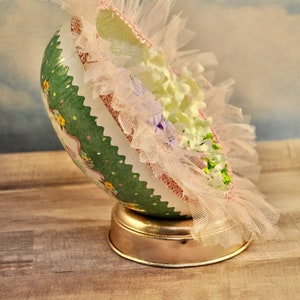 May include: A green and white decorated Easter egg with pink and white lace and flowers. The egg is on a gold stand.