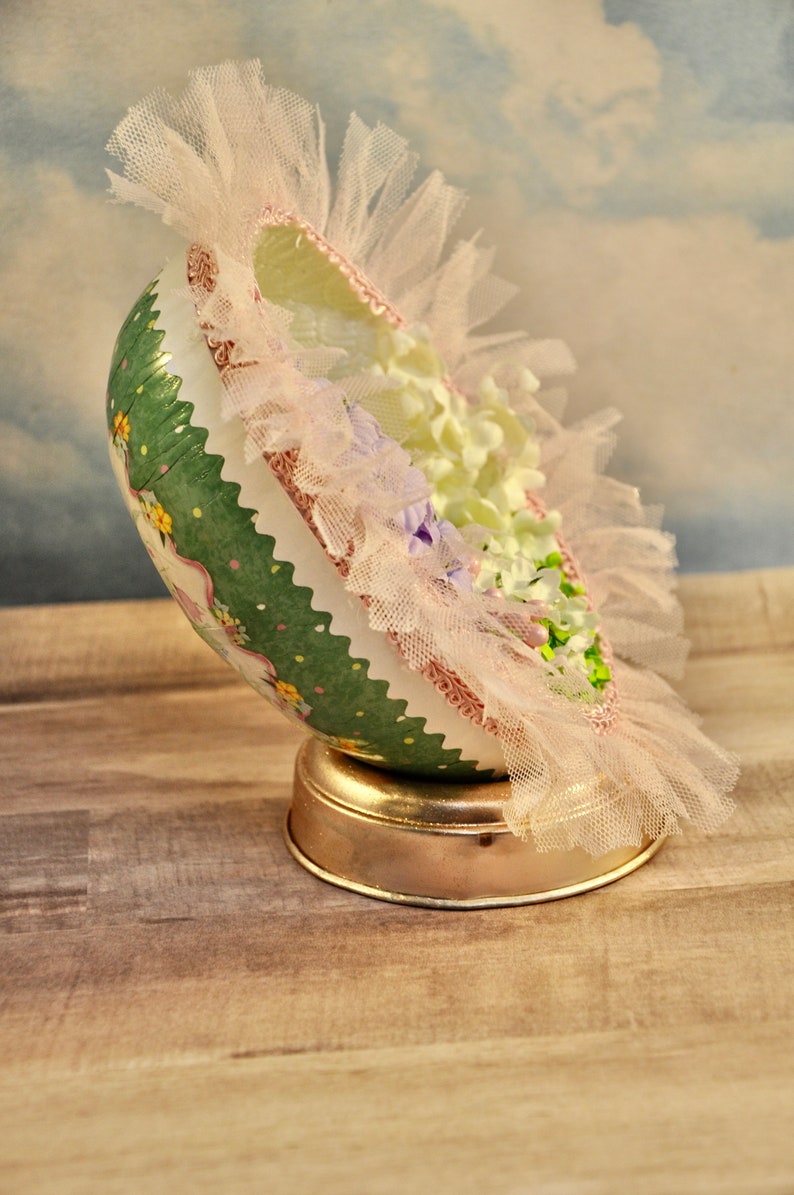 May include: A green and white decorated Easter egg with pink and white lace and flowers. The egg is on a gold stand.