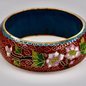 Pode incluir: Uma pulseira larga e redonda de cloisonné. A pulseira apresenta um fundo bordô escuro com detalhes dourados e desenhos florais em branco, verde e roxo. O interior é azul escuro e as bordas são adornadas com ouro e turquesa.