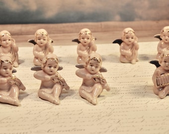 Puede incluir: Una colección de pequeñas figuras de querubines de porcelana. Los querubines están en varias poses, algunos tocando instrumentos musicales como violines y acordeones. Cada querubín tiene detalles dorados y alas de color gris oscuro. Las figuras están sobre una superficie de papel.