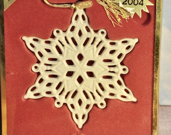 May include: A white porcelain snowflake ornament with a gold tassel. The ornament is in a red box with a gold border and a gold star with the text "NEW 2004". The box also has the text "2004 ANNUAL" and "LENOX".