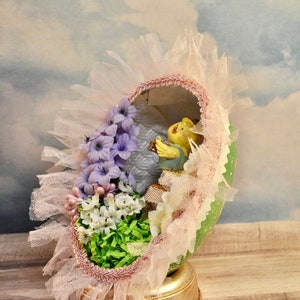 May include: A decorated Easter egg with a pink and white lace trim, purple and white flowers, and a small yellow figurine inside. The egg is sitting on a gold stand.