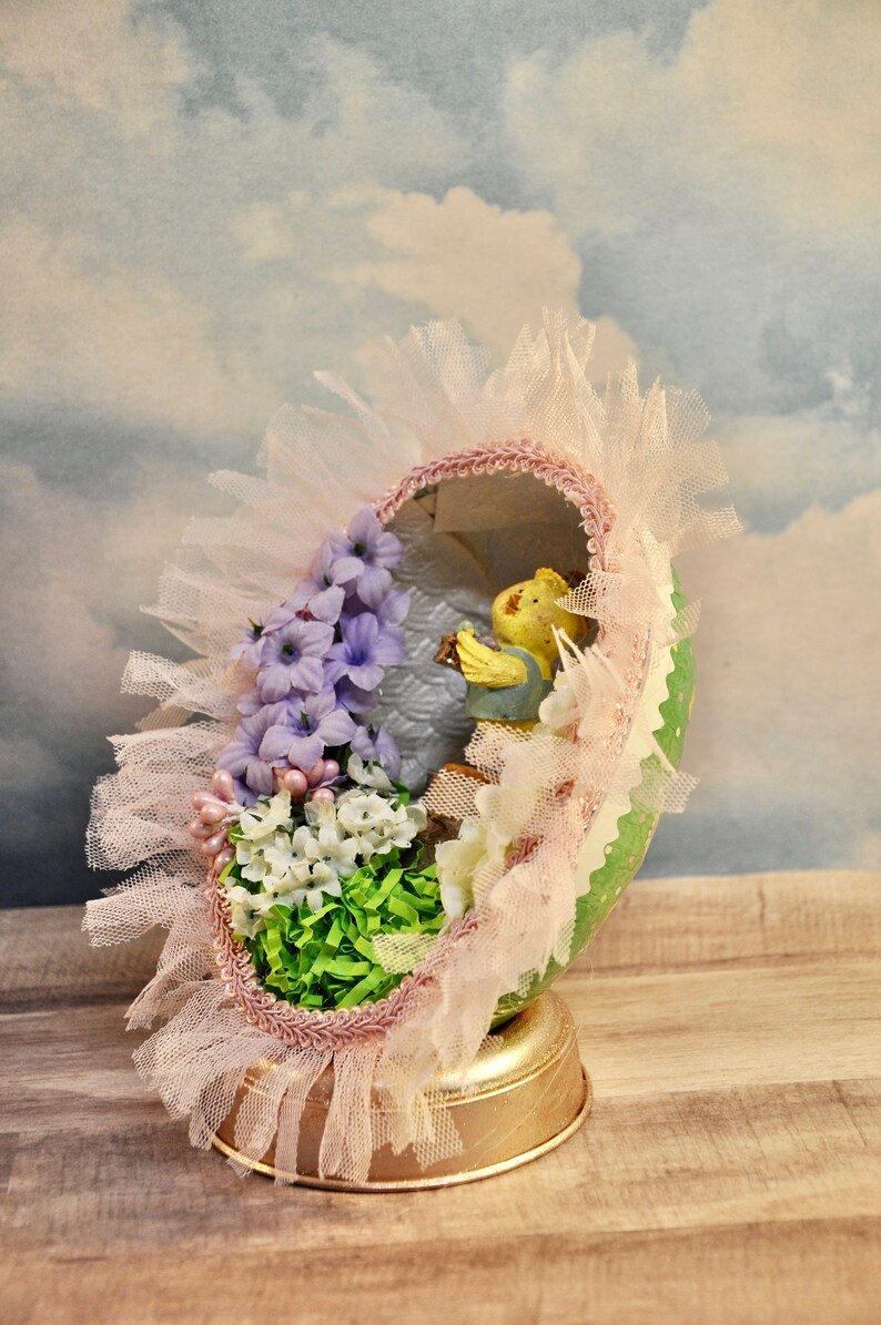 May include: A decorated Easter egg with a pink and white lace trim, purple and white flowers, and a small yellow figurine inside. The egg is sitting on a gold stand.