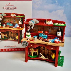May include: A red wooden workbench ornament with a variety of tools and toys, including a toy train, a rocking horse, a penguin, a snowman, and a Santa mug. The ornament is decorated with a festive red and green color scheme and features the text "Santa's Workbench".