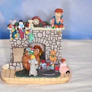 May include: A Christmas-themed decorative fireplace figurine. The stone-textured fireplace features a clock, stockings, and miniature figures, including a snowman, teddy bears, and a seated man. A train and other toys are also present.