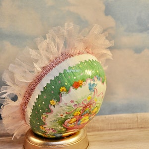 May include: A green and white Easter egg with a pink tulle ruffle and a floral design. The egg is sitting on a gold stand.